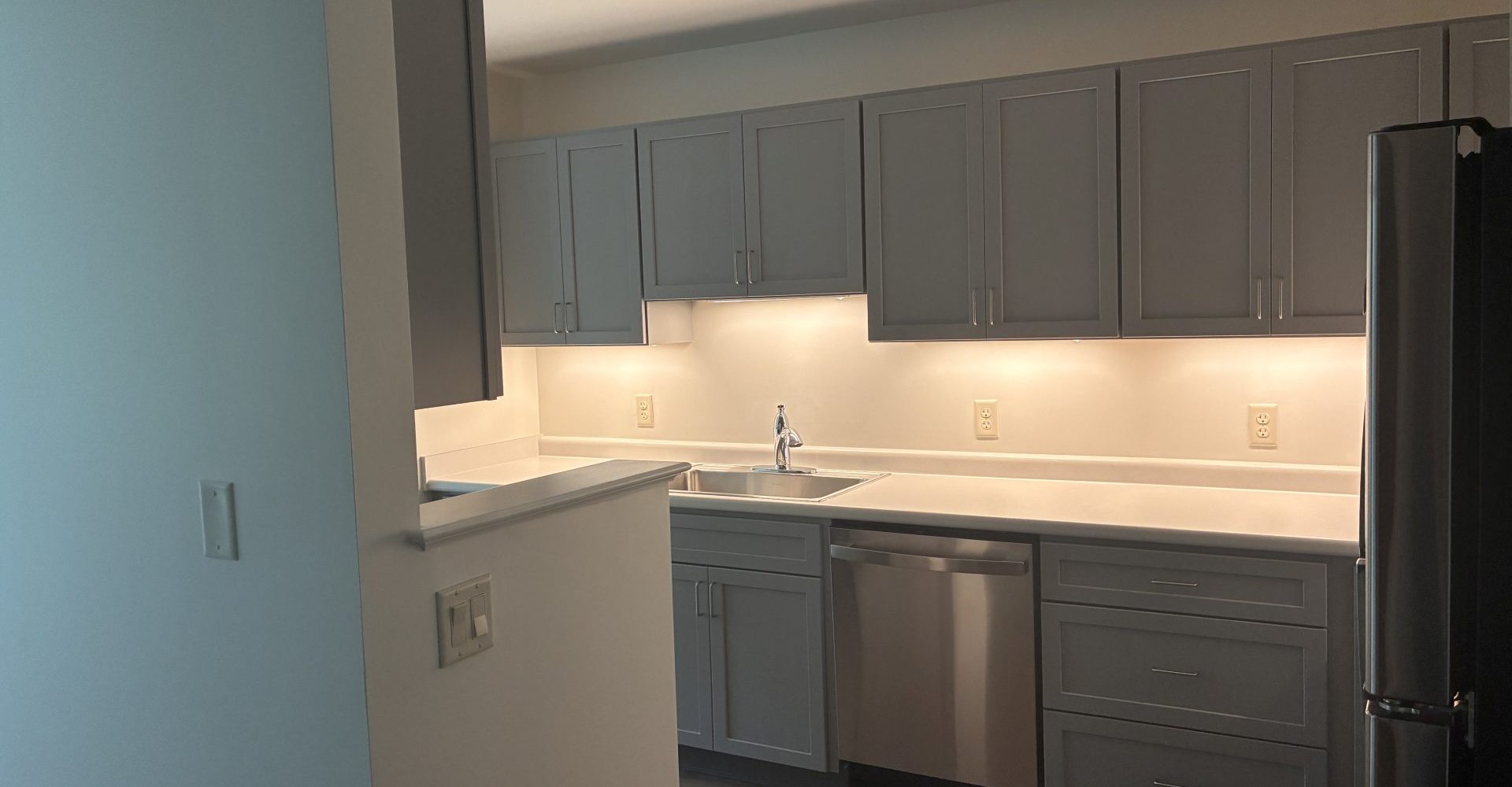 Kitchen with gray cabinets, white countertops, stainless steel dishwasher and refrigerator, and under-cabinet lighting.