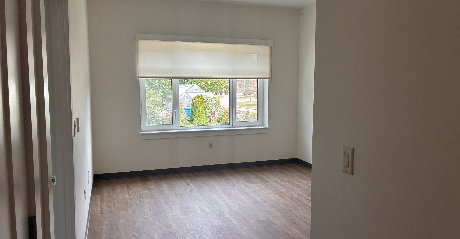 Empty senior apartment bedroom with wood-look flooring, a large window overlooking trees, and a closet.
