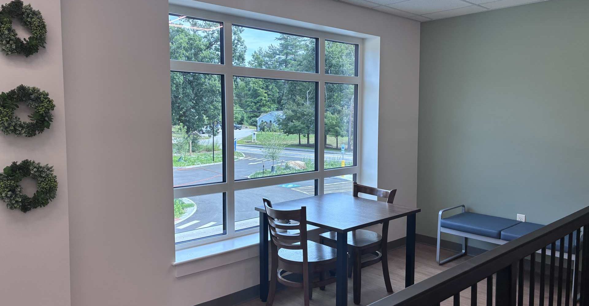 A small seating area with a table, chairs, and bench near a large window overlooking a landscaped street view.