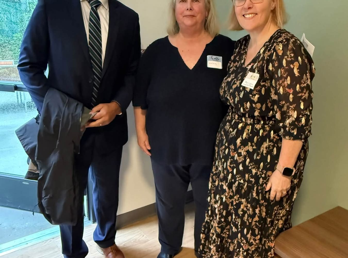Man in a navy suit stands with two women wearing name tags at an indoor community gathering