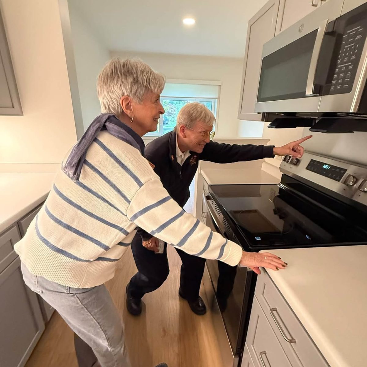 Two seniors smile while pointing at the new stove and microwave in a bright apartment kitchen