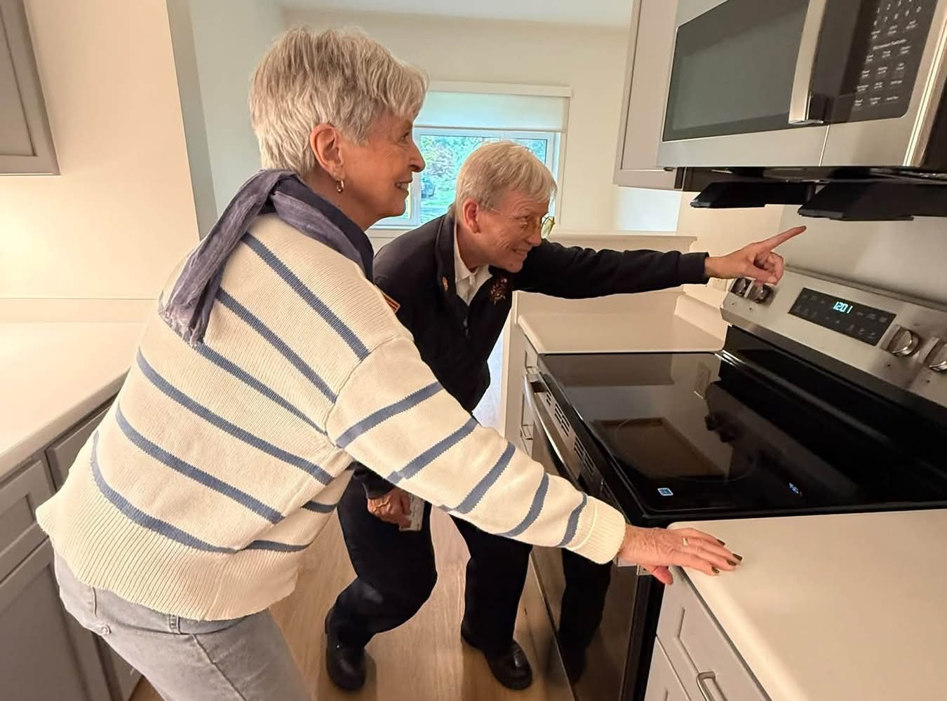 Two seniors smile while pointing at the new stove and microwave in a bright apartment kitchen