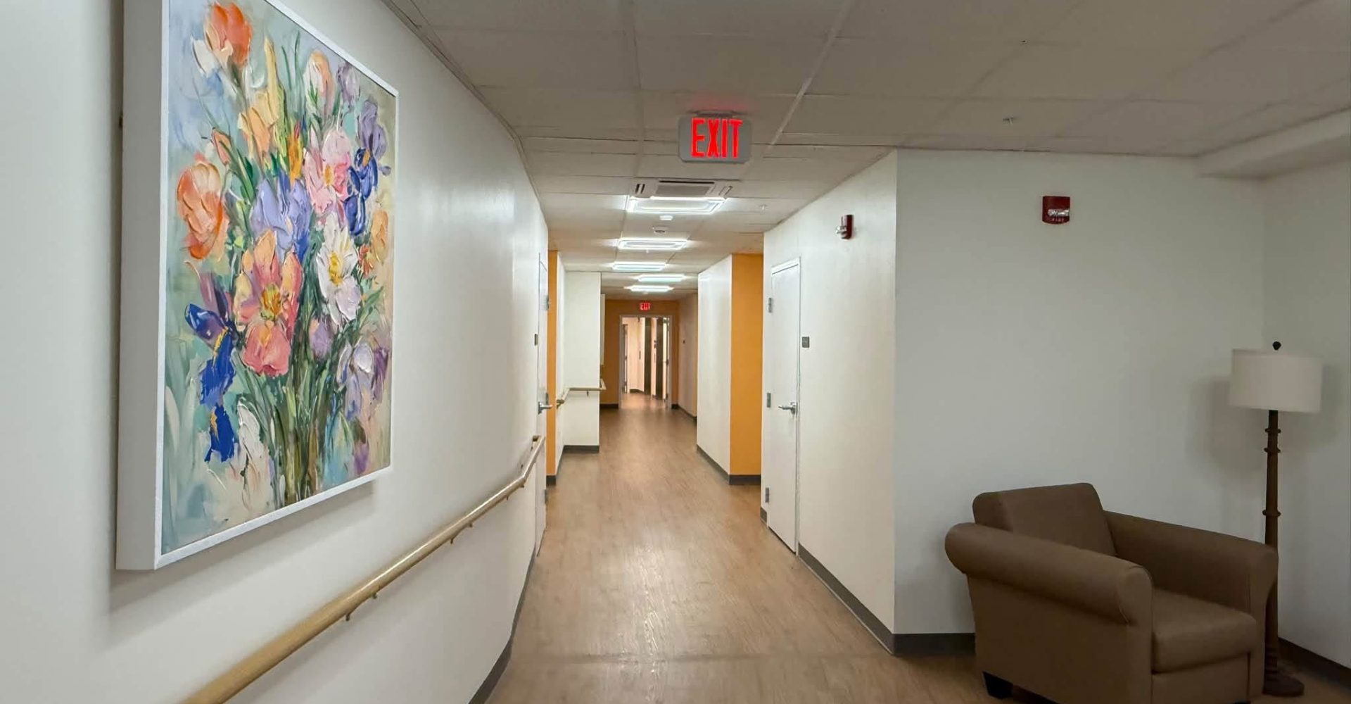 Hallway with handrails, a floral painting, seating area, and wood-look flooring leading to resident doors