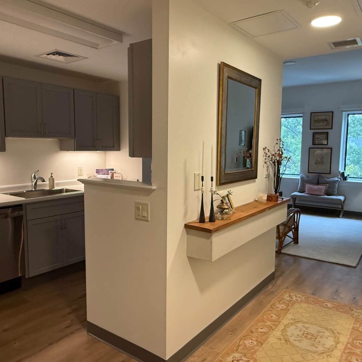 Open senior apartment layout with gray kitchen cabinets, a wall mirror console, and a living room with windows beyond