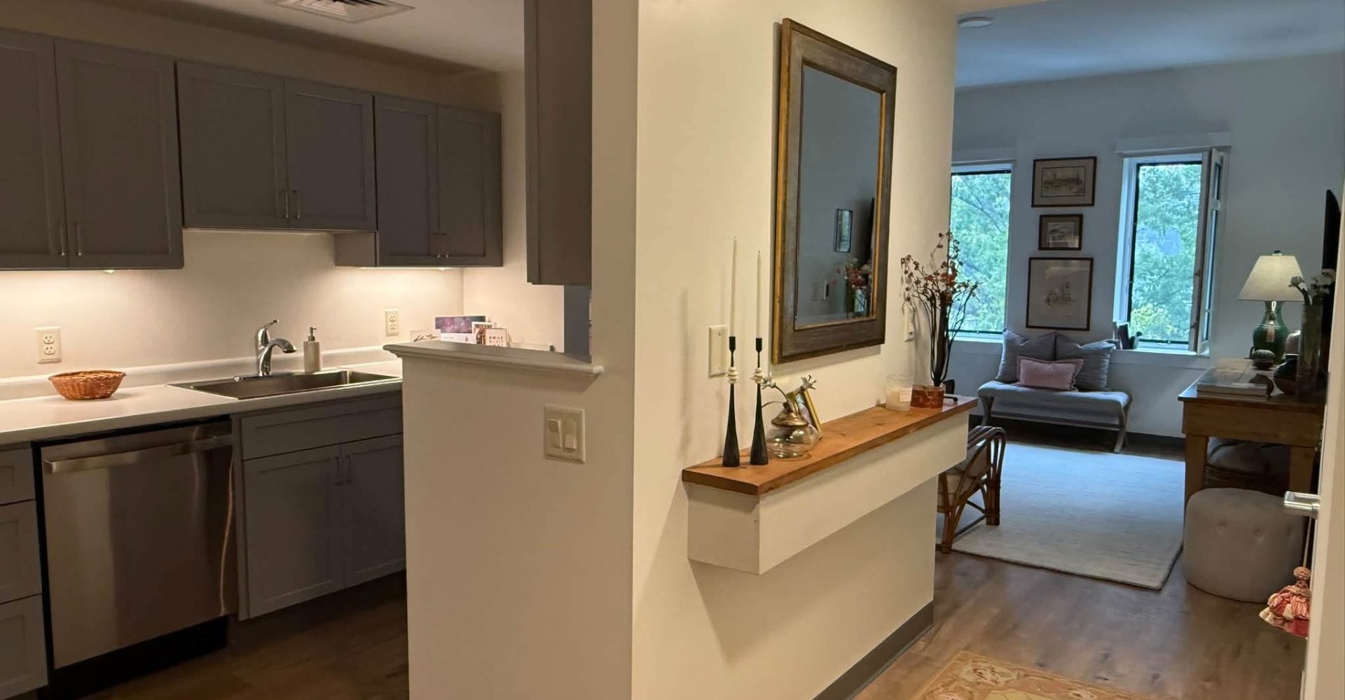 Open senior apartment layout with gray kitchen cabinets, a wall mirror console, and a living room with windows beyond